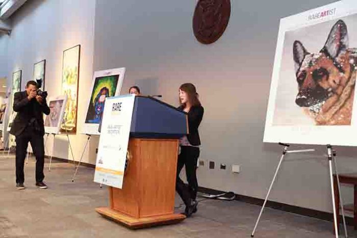 Elizabeth stands at a podium speaking. Award winning art is on easels next to the podium.