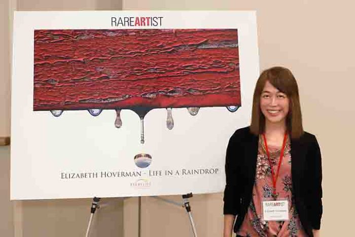 Elizabeth Hoverman is standing next to a poster on an easel of her award winning photograph, Life In a Raindrop. 