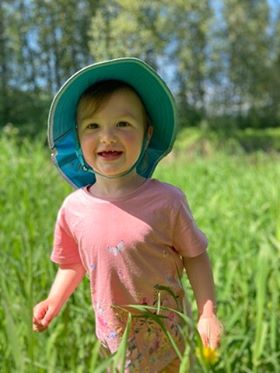 Clara is standing in a field of tall grass wearing a pink shirt and a blue sun hat.