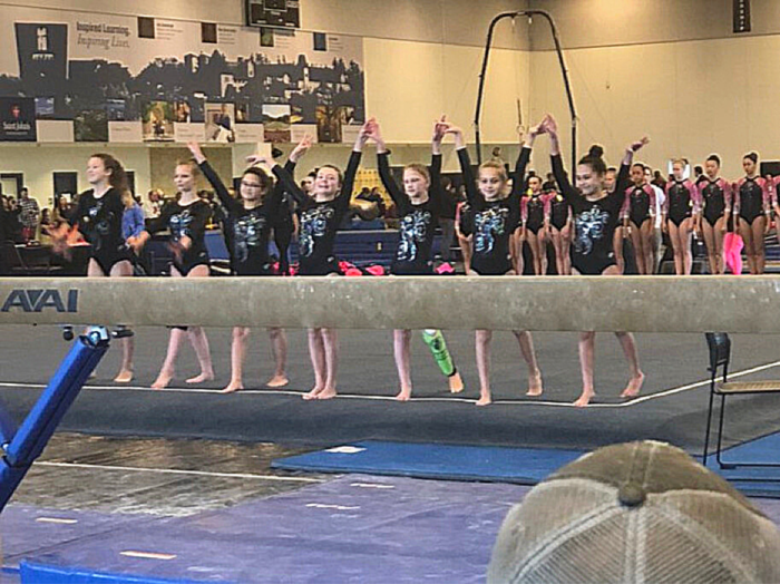 Jordan stands with 6 gymnasts from her team, with hands in the air at a gymnastic competition. They are behind a balance beam.