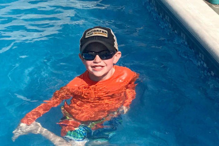 Jamistyn is in a swimming pool wearing a blue baseball hat, black sunglasses and an orange shirt. He's smiling and holding his right foot.