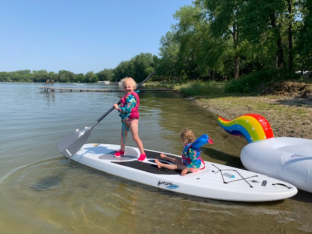 Ava is standing on a paddleboard on a lake wearing a life jacket. She's holding a paddle. Her little sister is sitting on the paddleboard, wearing a life jacket.