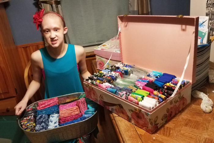 A young woman with a blue tank top and pink flower in her hair holds a basket in her lap filled with folded bandanas. On the table next to her is another box filled with folded bandanas of various colors and patterns.