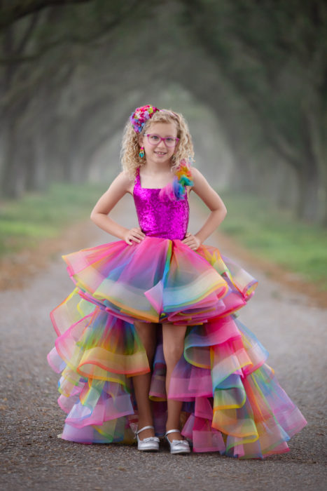 Lily is standing outside and wearing a rainbow formal dress with a rainbow colored bow in her hair.