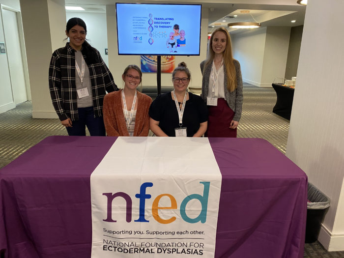 Four women pose behind a table with and NFED table cloth on it and the International Ectodermal Dysplasias Conference logo on a screen behind them.