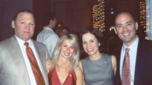 Four adults stand arm in arm. There are men on the outside wearing jacket and ties and the ladies are wearing cocktail dresses. They are attending the NFED Halloween Bash.