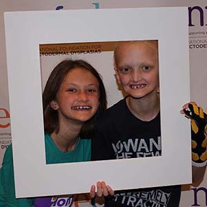 A girl and Keegan have their faces inside a poster frame they are holding up with their hands.