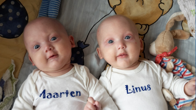 Twin baby boys affected by XLHED lay on a baby blanket with a stuffed bunny. The baby on the left wears a white onesie with Maarten printed on it. The other baby wears a white onesie with Linus printed on it. 