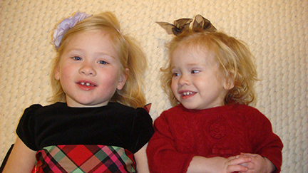 Two toddler girls in Christmas dresses are laying on the floor side by side. Each has blonde hair and a hair bow. The girl on the left is showing just a few teeth.