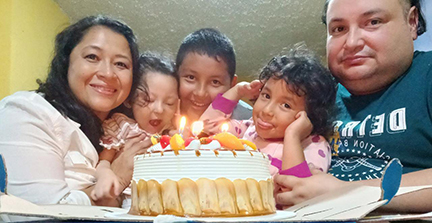 A mom and dad with their baby daughter, a son and another daughter are shown with a birthday cake that has three candles on it.