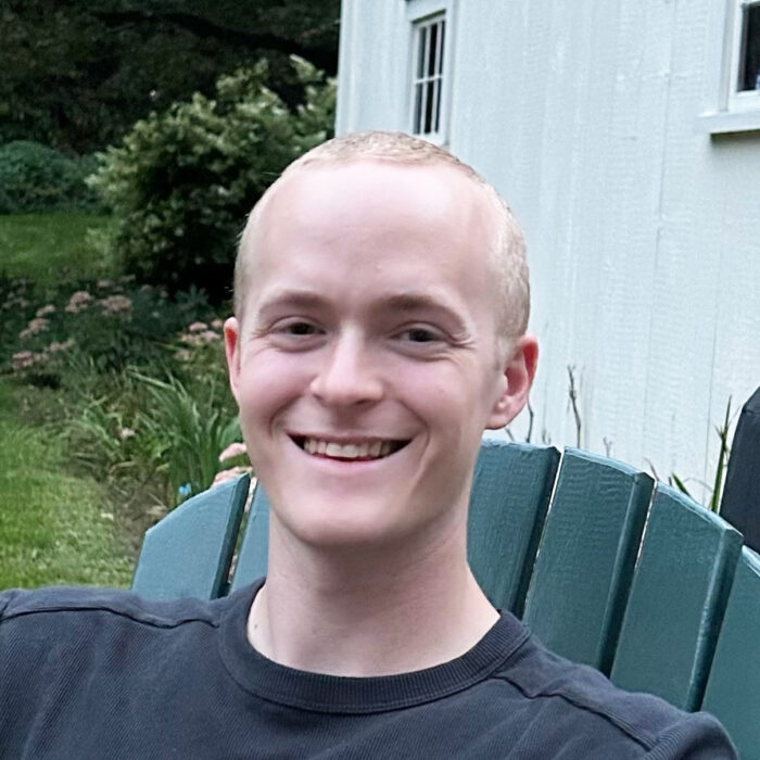 This is a headshot of Soren who is wearing a black tshirt and sitting in a green Adirondack chair outside of a while house.