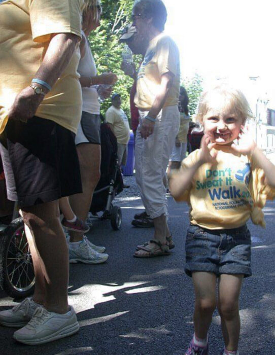 Nicole is a little girl wearing a Don't  Sweat It Walk shirt with blue jean shorts. She has white blonde hair. Adults attending the walk stand in the background.