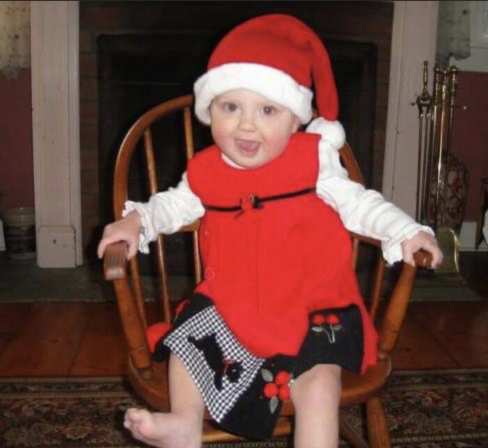 A toddler is wearing a Santa Hat, white shirt and a red jumper as she sits on a wooden chair.
