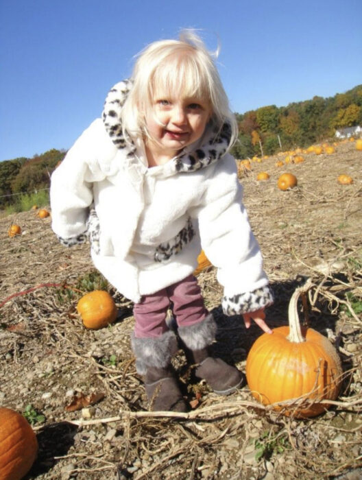 Nicole is a little girl in a white coat with black boots and she's picking pumpkins in a patch. 