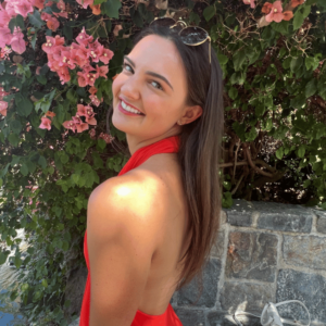 Lexie is wearing a red halter dress with sunglasses on the top of her head. She's smiling to show her beautiful teeth. There's a bush in the back with pink flowers.