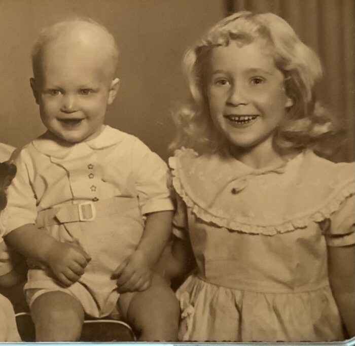John is a toddler with sparse hair and is sitting next to his older sister. She has blonde hair and is wearing a dress. The picture is in sepia.