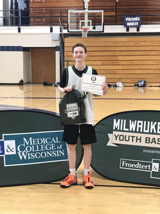A teenage boy is standing on a basketball court and holding a backpack and certificate.
