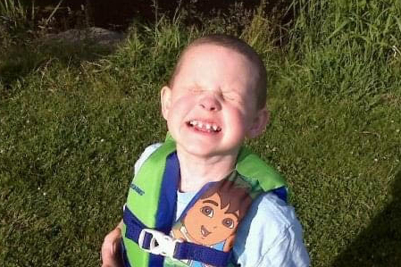 A young boy is wearing a life-jacket outside over a blue t-shirt. He's smiling and has missing teeth and teeth that are widely spaced apart.
