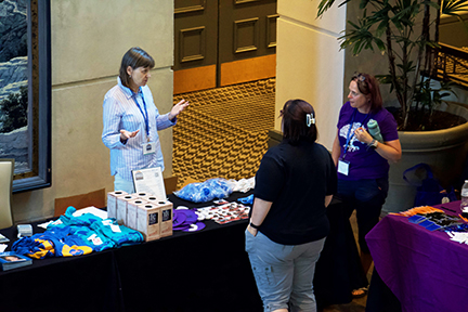 A woman with a lanyard around her neck stands behind a table selling NFED shirts, fans, bracelets and other items. Two women stand in front of her talking to her.