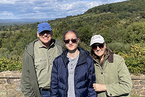 Marianne, her husband and son stand in front of hills. Each are wearing sunglasses and coats.