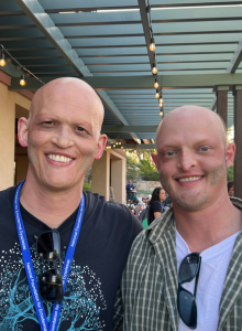 Two bald men stand arm in arm outside.