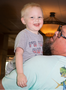 A man wearing glasses and a goatee lifts up his toddler son. The boy has sparse hair and missing teeth.
