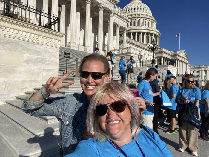 Two women smiling and wearing sunglasses stand on the steps of the U.S. Capitol with other advocates in the background.