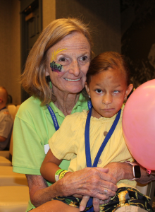 A woman with NFED painted on her face holds a young girl who is affected by Goltz syndrome. The little girl is holding a pink balloon and is wearing a yellow shirt.