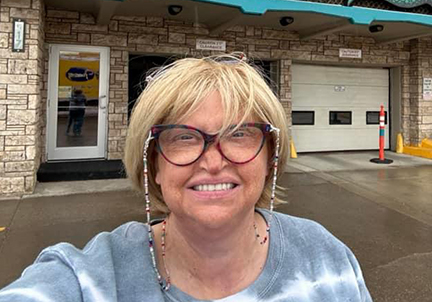 A woman with blonde  hair and wearing glasses is standing in front of a store. 