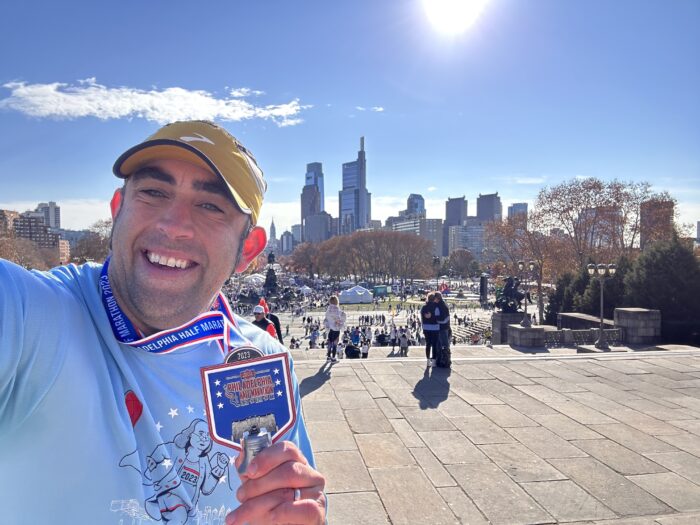 Greg is wearing a yellow hat, and a blue shirt and medal around his neck. He's standing outside with a crowd and the Philadelphia skyline in the backgroun.