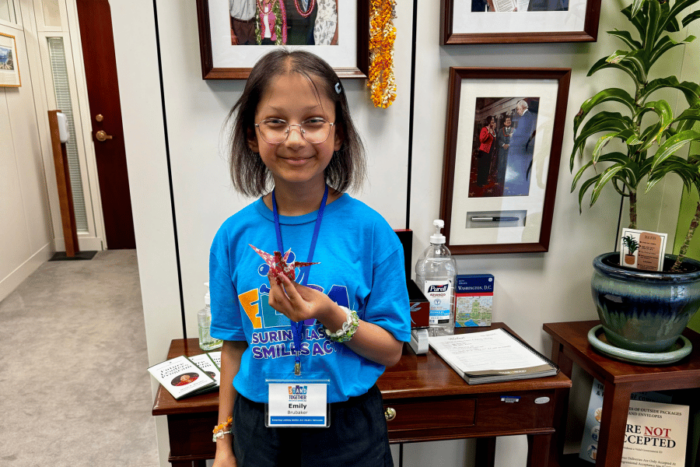 Emily is holding an origami crane at her legislator's office. Emily and parents share her ectodermal dysplasia journey with Sen. Murkowski's staff member.