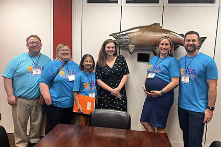 Five ELSA advocates stand with a senator's staffer in front of a large mounted fish on the wall. 