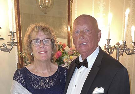 Liza and Frank are in formal wear standing in a ballroom in front of a mirror and flowers.