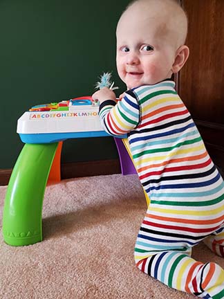 Jacki is wearing a striped onesie. He's kneeling, playing with a toy and smiling.