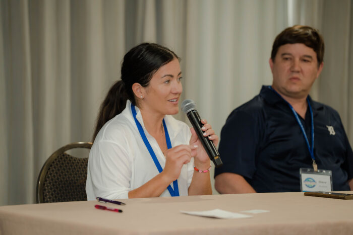 A woman with brown hair is speaking into a microphone while sitting at a table. A man to her left is watching and listening. Both are wearing conference lanyards.