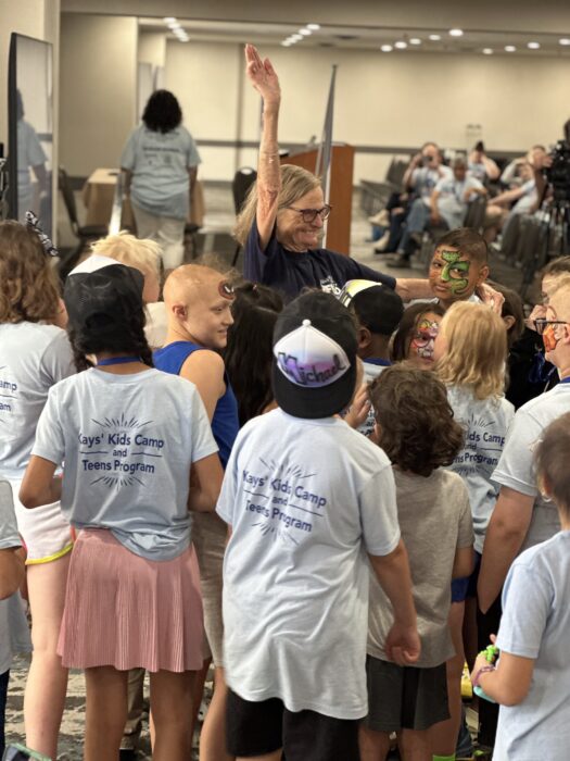 A woman stands in the middle of a crowd of children, smiling with her arm in the air.