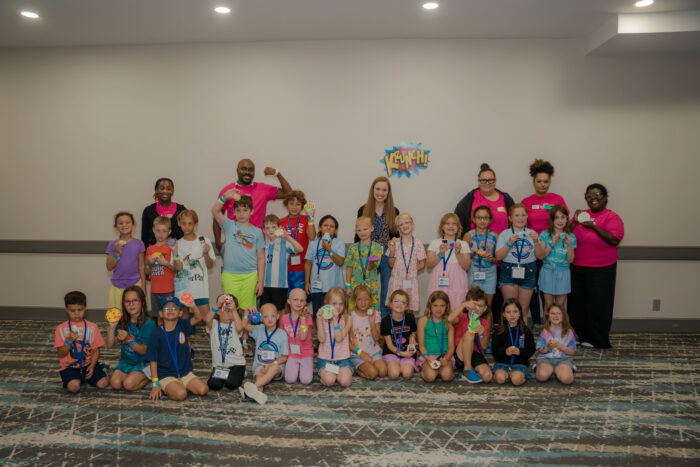 A large group of children line up for a photo with adult chaperones in a hotel conference room.