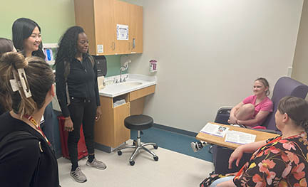 Two people sit in chairs in a medical examination room while four medical professionals ask them questions.