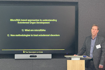 Dr. Amendt is standing at a speaker's podium wearing a suit coat. His presentation is on the screen in the backrground.
