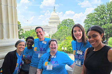 Six people stand on a balcony with trees and the U.S. Capitol building standing in the background.