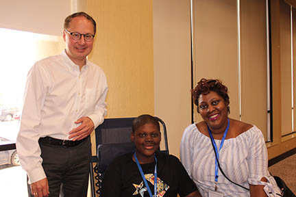 Dr. Stanford is standing next to a young man affected by ectodermal dysplasia who is sitting in a reclined chair with his mother on the right.