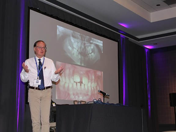 Dr. Stanford is standing in front of a large screen where intraoral photos are being shown. He's giving a lecture.