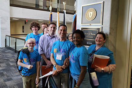 Nine people stand in a hallway in the Senate building outside of Senator Warren's office. Four flags on poles stand behind them.