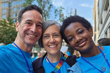 The three people stand together outside with their heads touching. All are wearing the same blue t-shirt. 