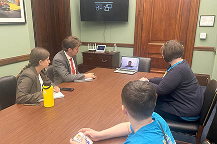 Four people sit around a conference table in an office looking at a laptop where a man is on screen.