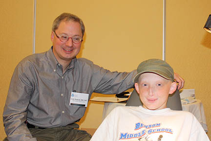 Clark sits next to a young man affected by XLHRF who is laying in a dental chair.