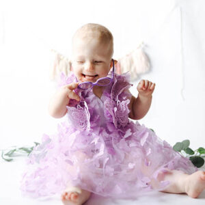 A baby girl is sitting up and wearing a purple dress with lots of ruffles.