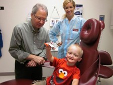 Dr. Stanford is standing next to a young boy sitting in a dental chair. The boy has XLHED, is wearing a red Elmo shirt and holding up his dentures. A dental hygienist stands in the background.