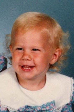 Lindsey as a young girl wearing a blue dress with a white collar. She has blond, curly hair.