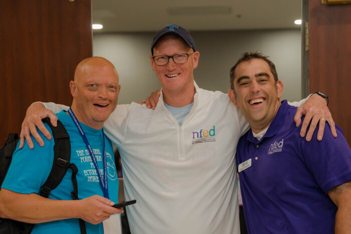Karl has his arms around Charley and Greg. They are standing in a conference space.